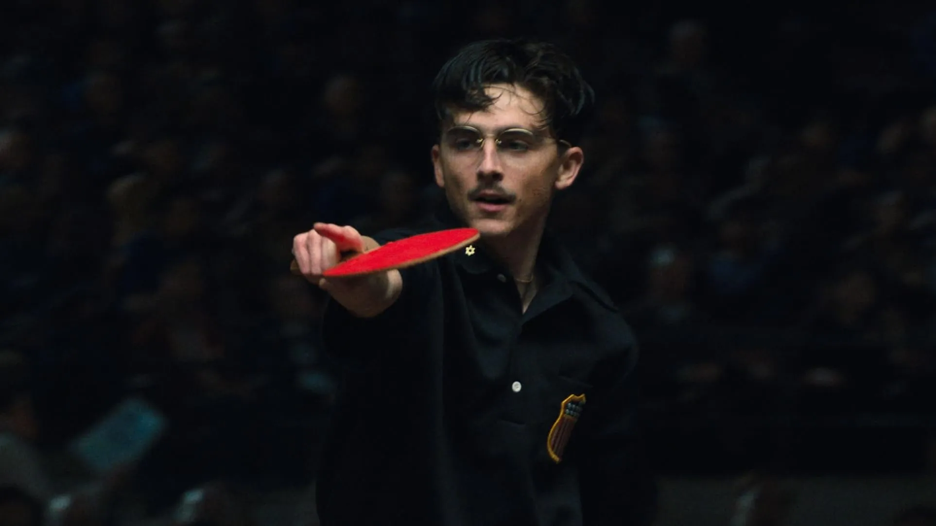 Timothée Chalamet as Marty Mauser, pointing a red ping-pong paddle in front of a dark crowd