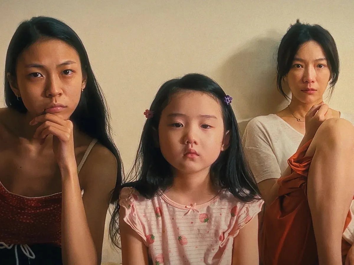 The older sister, the young girl and their mother sit side by side against a wall, looking straight at the camera